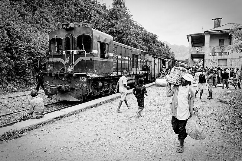 Locomotive du train FCE à l’arrêt en gare de Tolongoina, avec des voyageurs et porteurs en pleine activité sur le quai